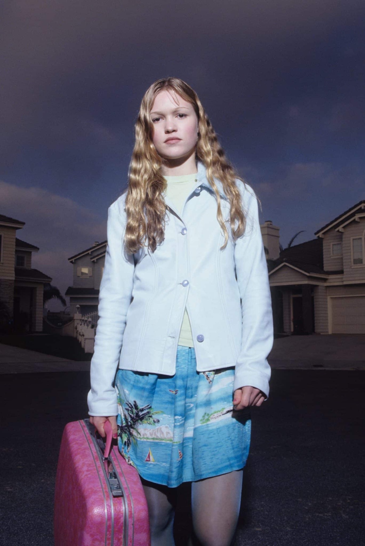 Ellie standing alone in the middle of the road at dusk in a
               white jacket and a seaside-print skirt, a pink suitcase in one
               hand, the identical tile-roofed houses of Casa Del Norte receding
               behind her under a bruised sky.