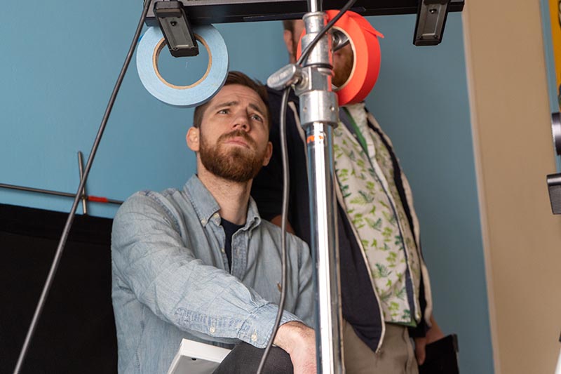 Image of Curtis Clark, standing in a film studio behind a stand with 2 rolls of duct tape hanging from it. 