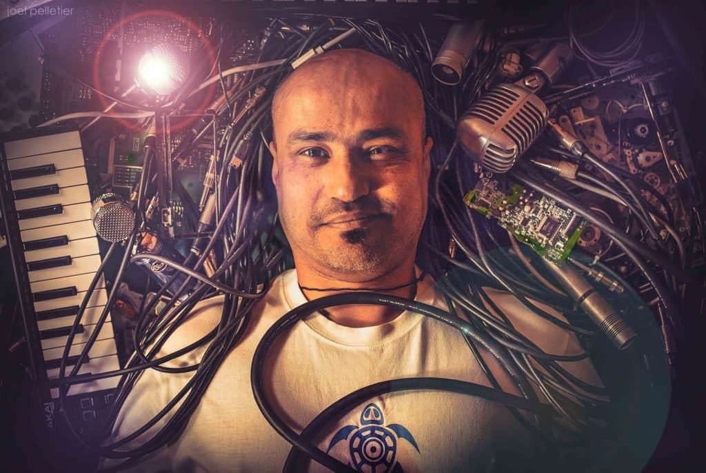 image-of-Adham-Shaikh-music-producer-surrounded-by-instrument-cables-microphones-keyboard-MIDI-controller-rackmount-effects-systems-and-other-music-production-tools