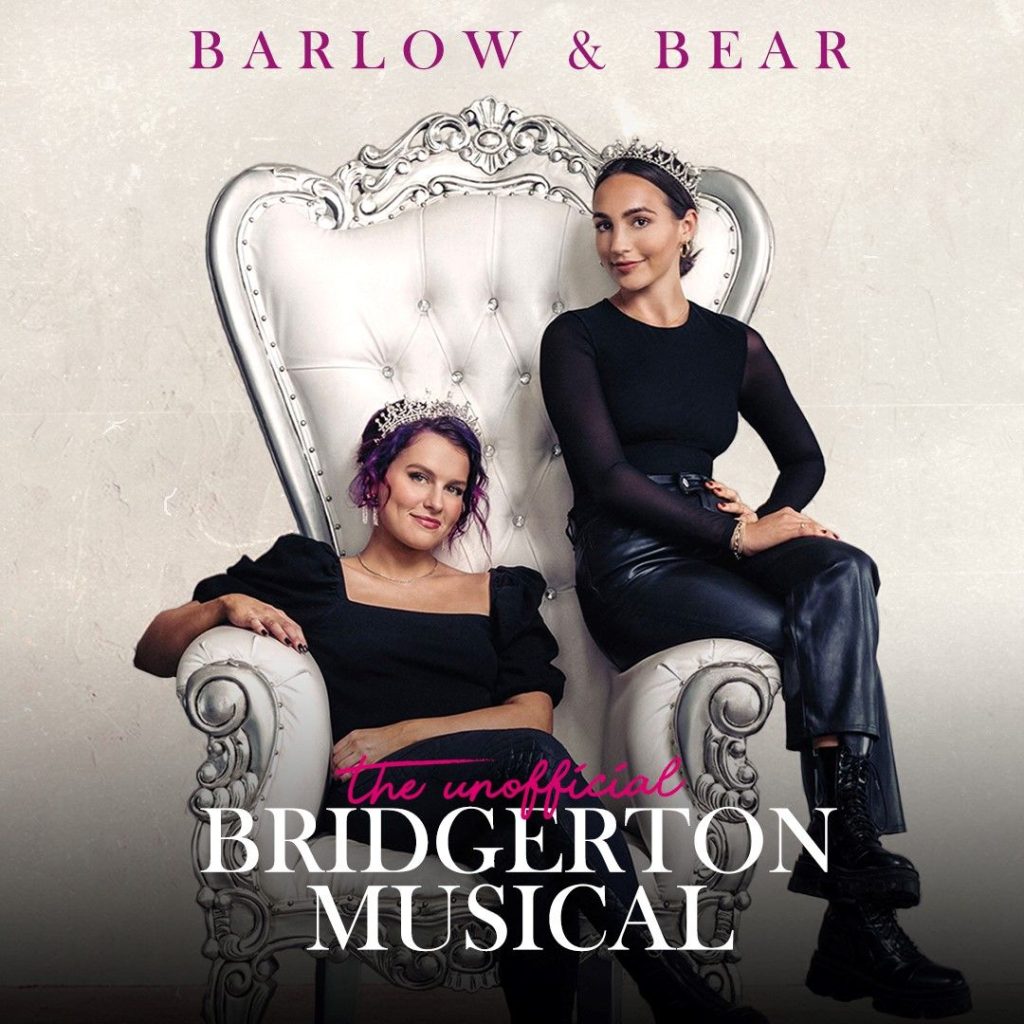 Barlow and Bear sit on a white and silver throne together wearing tiara crowns in a publicity photo for the Unofficial Bridgerton Musical