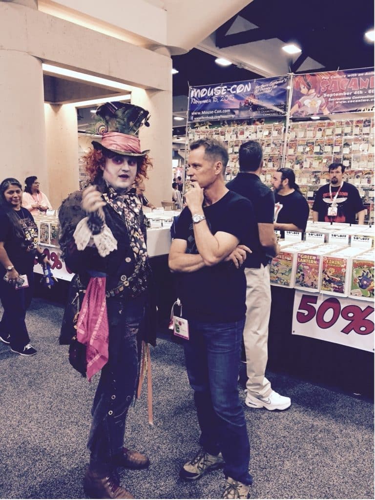 In the image, Chad Evett, dressed as the Mad Hatter, and Frank Beddor, a man in a casual outfit, are standing next to each other, discussing something intently.