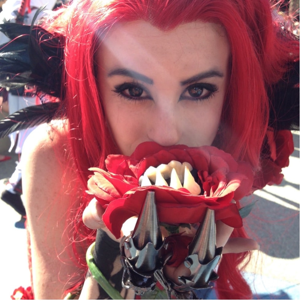 Queen Redd cosplay designed by Chad Evett features a model with red hair and details like a killer flower, metal guarded gloves, and black feathers for her hair.