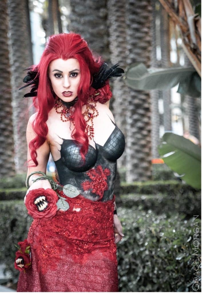 Queen Redd cosplay designed by Chad Evett features a model with red hair and details like a killer flower, metal guarded gloves, and black feathers for her hair removes her top to show a body paint undergarment.