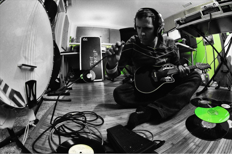 Black-and-white-image-of-music-artist-Phontaine-playing-a-mandolin-on-the-hardwood-floor-of-a-music-studio-with-various-instruments-vinyl-LP-records-and-recording-gear-surrounding-him