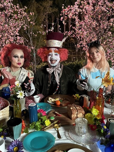 A group of people sitting at a table with food and flowers, dressed like characters from Lewis Carroll's "Alice's Adventures in Wonderland. Red Queen, Mad Hatter and Alice Liddell, herself.  For the rapper, Dax and hiis song: "Searching for a Reason". 