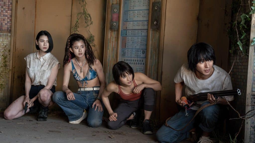 Image of the cast of the popular Netflix series: Alice in Borderland. The four of them are kneeling down and hiding in a Tokyo entryway, with the closest character holding a long, double-barrel rifle. 