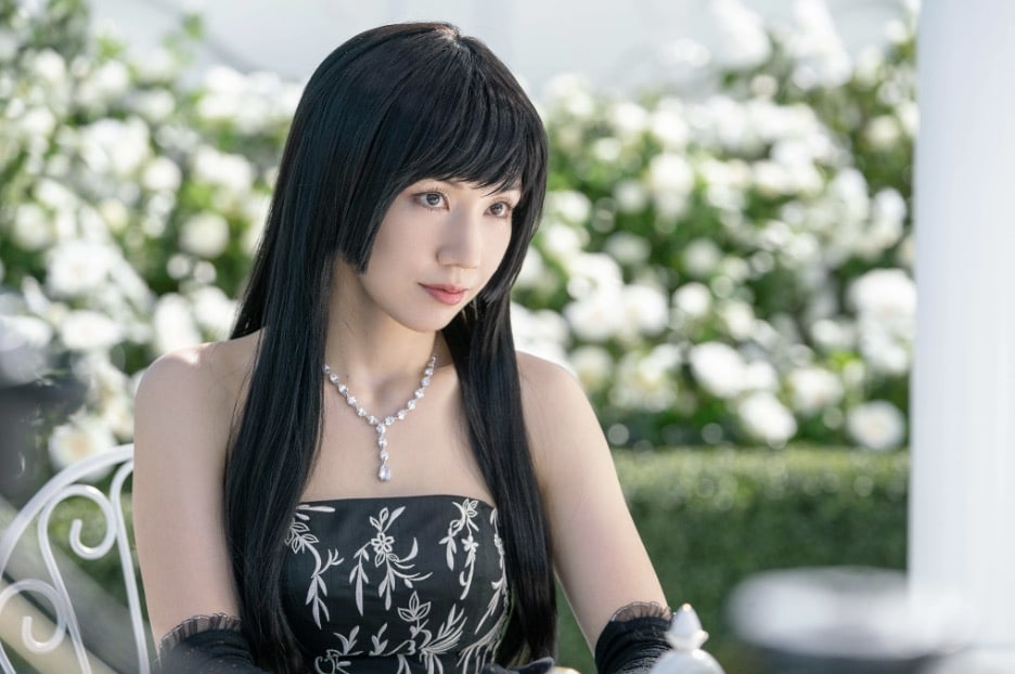 Image of actress Riisa Nana from Alice in Borderland, the popular Netflix series based on a Manga that is based off of Lewis Carroll's Alice in Wonderland. She is sitting in a garden tea party with white flowers, furniture and building structure, while wearing an elegant black dress with a white floral pattern. 