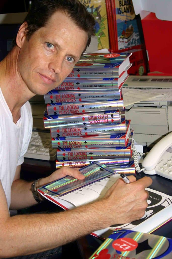 Author, Frank Beddor, signing copies of his first edition hardcover book: The Looking Glass Wars, sitting at a desk with a stack of books, a telephone and a fax machine. 