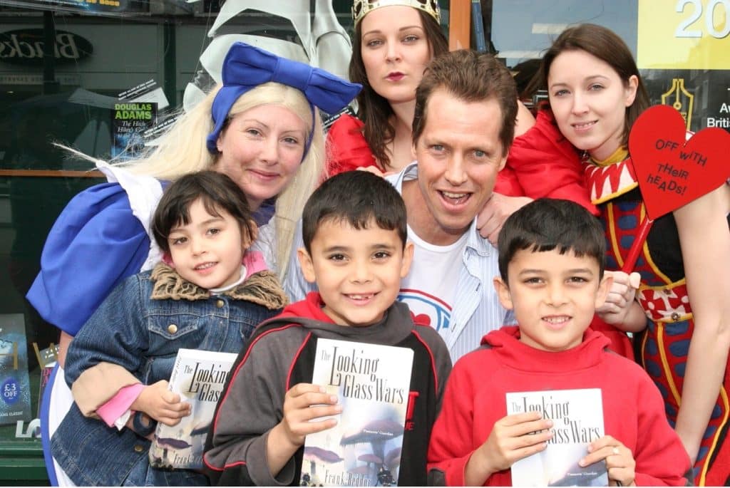 Author, Frank Beddor, with a group of people, posing for a photo. He is joined by characters from Lewis Carroll's Alice in Wonderland, and some kids holding up copies of his book, The Looking Glass Wars. 