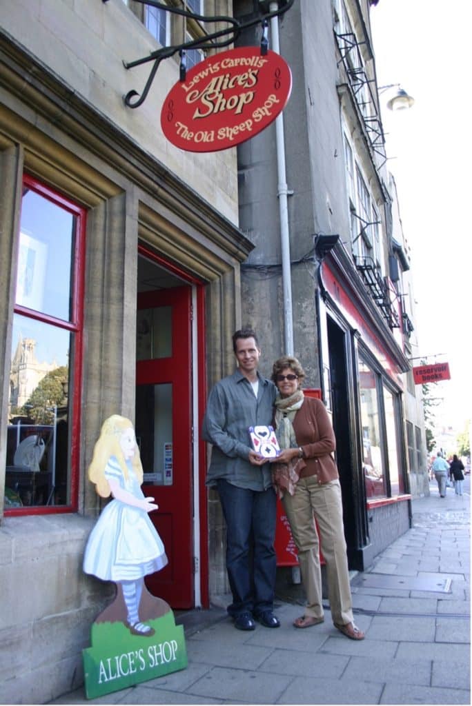 Frank Beddor and his agent, Barbara Marshall, standing in front of a store named: Lewis Carroll's Alice's Shop - The Old Sheep Shop. They are standing next to a life-sized cutout of Alice from Alice in Wonderland and holding up a copy of Frank's book: The Looking Glass Wars. 