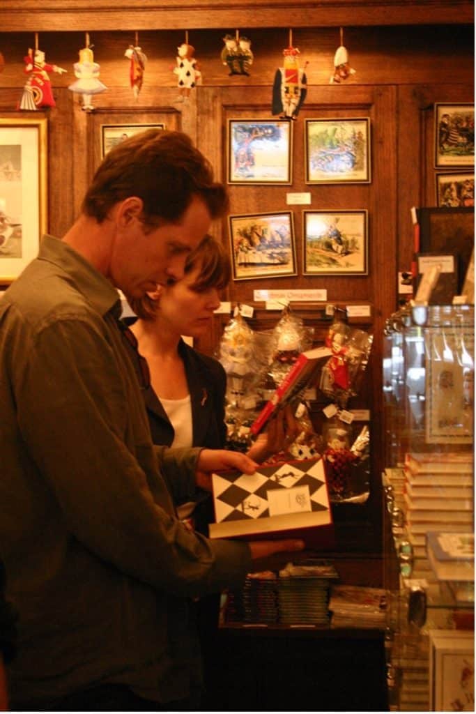 Frank Beddor and Cally Poplak, standing in an antique shop, looking at copies of Frank's book: The Looking Glass Wars, that is for sale in the shop. There are vintage Christmas decorations up around the mostly stained wood walled shop. 