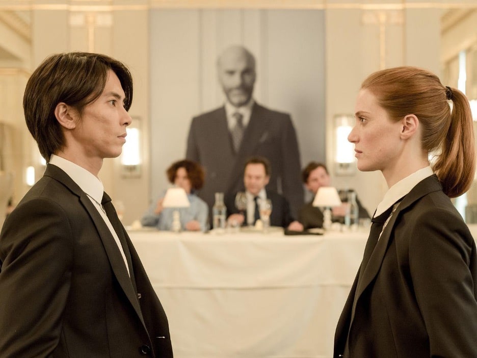 Image of 2 people staring each other down, with a table of 3 people looking onwards, judging them. A large portrait of some important man looms just out of focus in the background. From a TV series Drops of God