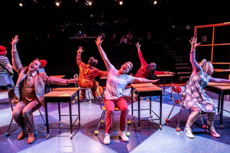 Bad Hat Theatre group sitting in school desks on stage for their performance of their unique adaptation of Lewis Carroll's Alice in Wonderland. Each actor is a student, raising their hands from their desks. 