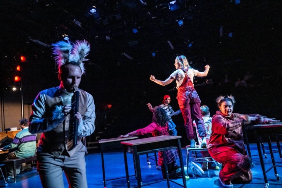 Photo from Bad Hat Theatre's production of Alice in Wonderland, during a musical number. The White Rabbit is playing the melodica, while Alice is standing on her desk, presumably singing a song. 