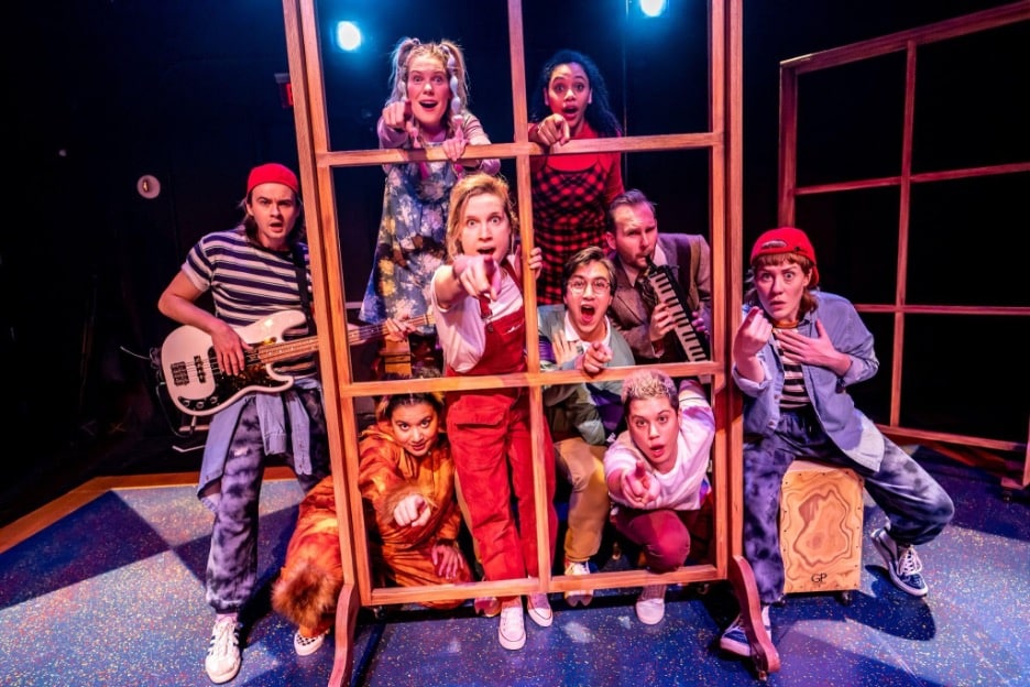 Photo of Bad Hats Theatre ensemble cast for their production of Alice in Wonderland. 9 cast members are standing on a stage, in costume, looking through window pane frames. 3 members of the cast are holding musical instruments, including a bass guitar, a melodica and a cajon. 