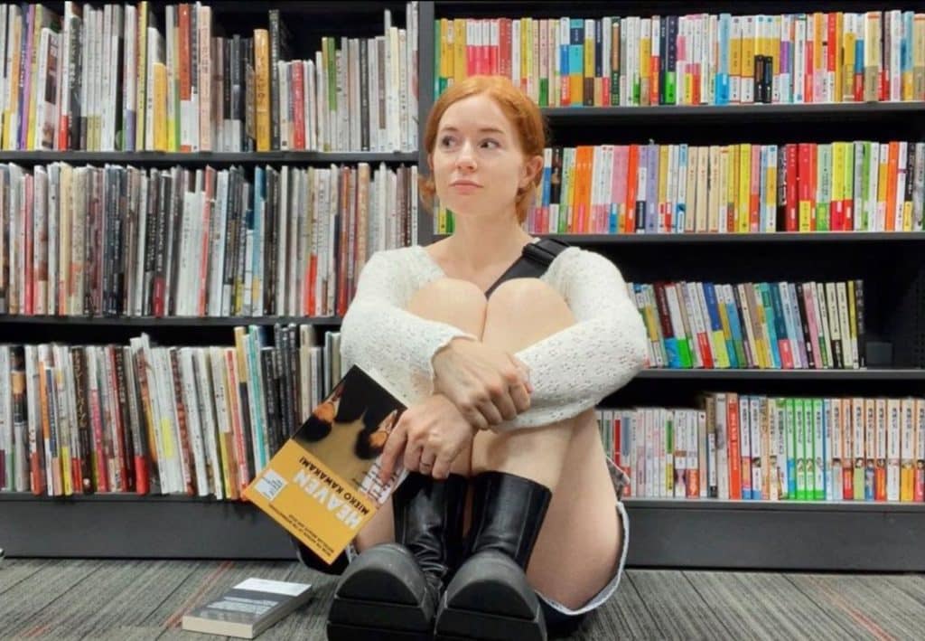 Image of Booktuber, Caricanread, who is sitting on the floor in front of a large bookshelf, that appears to be in a library. 