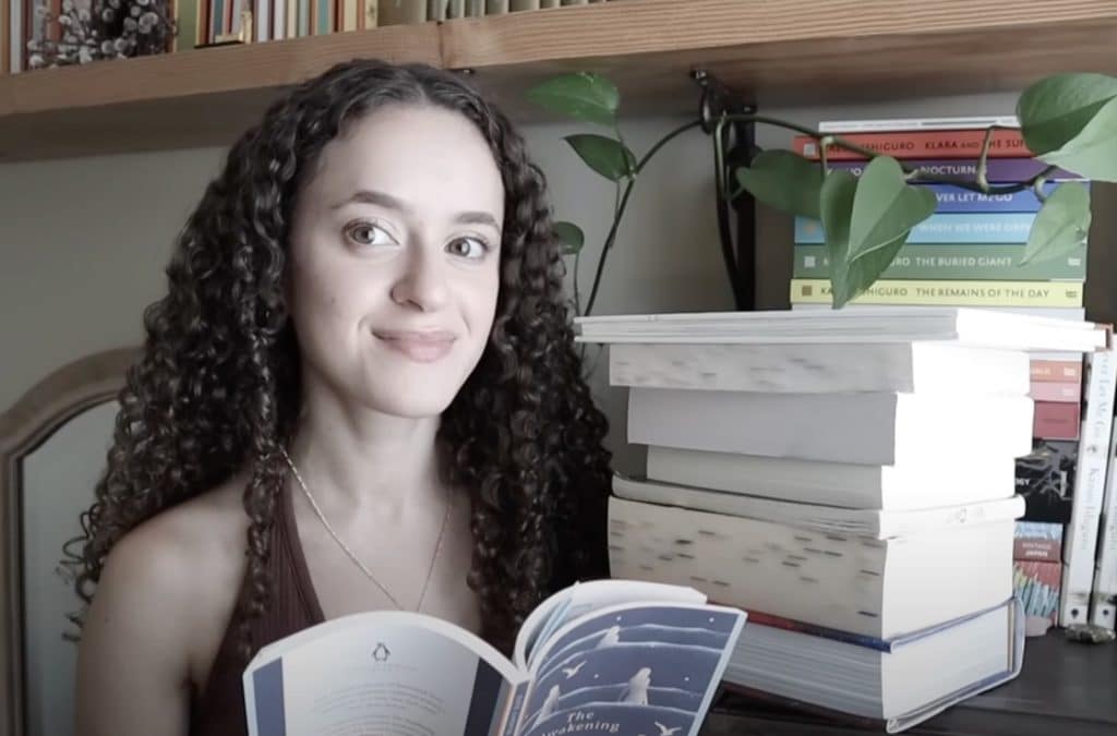 Image of Booktuber: CarolynMarieReads, reading a book called: "The Awakening". She is sitting at a small student's desk, with a lot of books stacked upon it.
