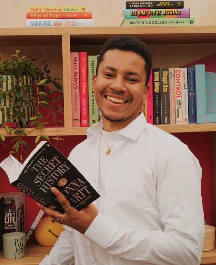 Image of Fictionalfates, a YouTube literary personality, or "Booktuber" who is smiling at the camera, while holding a book called "The Secret History", by Donna Tart He is wearing all white with a single gold chain. 