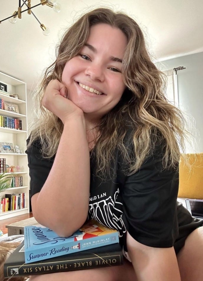 Booktuber, or Booktokker, HaileyinBookland sitting on a stack of books smiling for the camera. 
