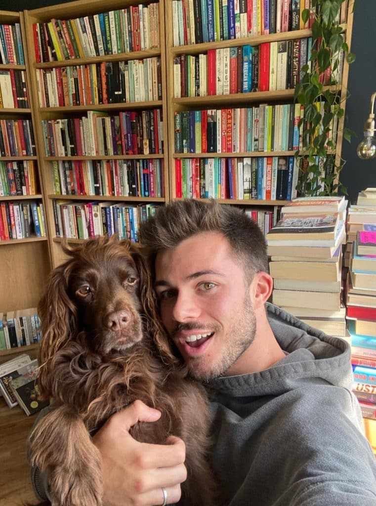 Image of literary content creator: Jack_Edwards who is holding a cute brown dog in front of a bookshelf and large stack of books on the floor. 