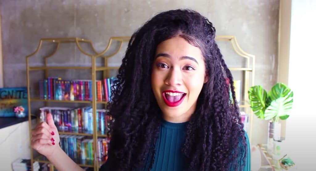 Booktuber NayaReadsandSmiles, with long, dark curly hair and red lipstick. She is wearing a green sweater and sitting in front of a large, gold bookshelf with a lot of books she has read. 

