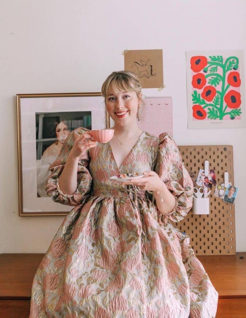 Booktuber: PeruseProject, in a dress holding a cup of tea and a small plate. She is standing in front of a wall with some art, a picture of green, red and black flowers, a painting of a woman and a bulletin board. 
