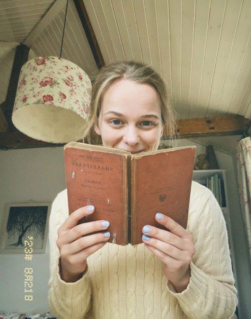 Image of YouTube literary content creator, Ruby Granger holding a book by William Shakespeare. She is wearing an off white sweater, sitting in an attic, where everything looks vintage. 
