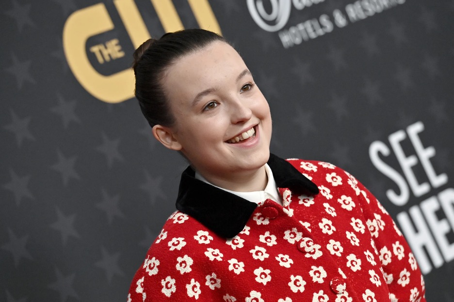 Image of Bella Ramsey, from The Last of Us and Game of Thrones fame. Here, she is smiling at the camera on the red carpet from a recent Hollywood premiere, wearing a red jacket with black collar and white floral pattern. 