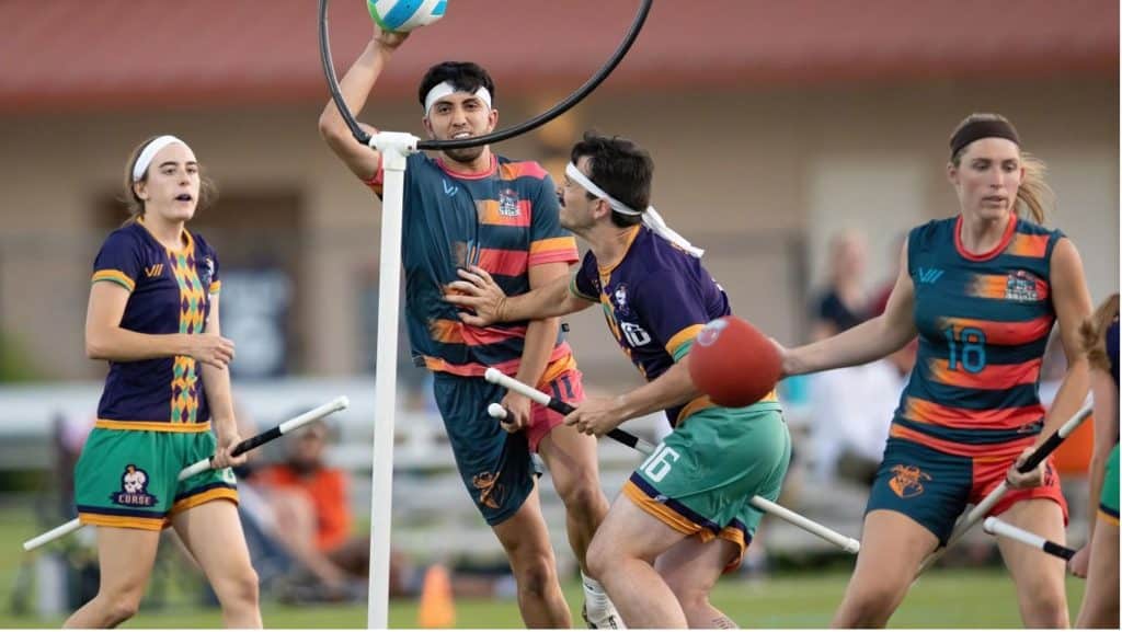 A group of people playing Quidditch, wearing outfits that look strikingly like rugby players. Here two players are riding broomsticks, and throwing a ball through a hoop, while two other players look on in amazement. 