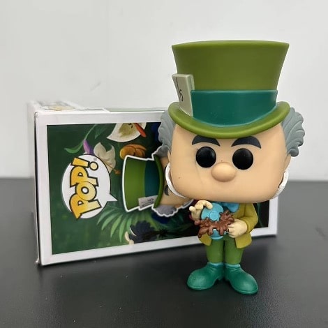 A toy figurine by Funk Pop of the Mad Hatter from Disney's Alice in Wonderland, wearing a green jacket, pants, shoes and hat. The pop figure is standing outside of its box, holding an overflowing cup of tea. 