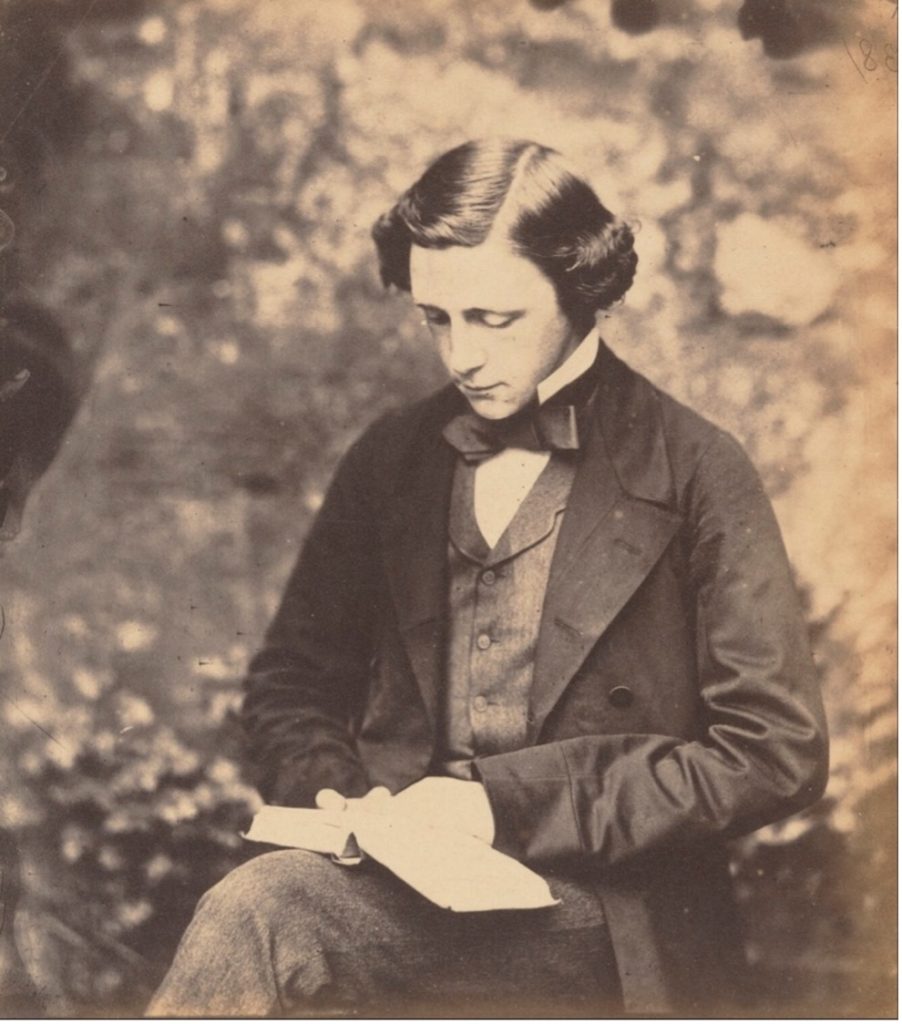 Old, black & white photograph of a young Lewis Carroll, wearing a suit  and reading a book in the forest. 
