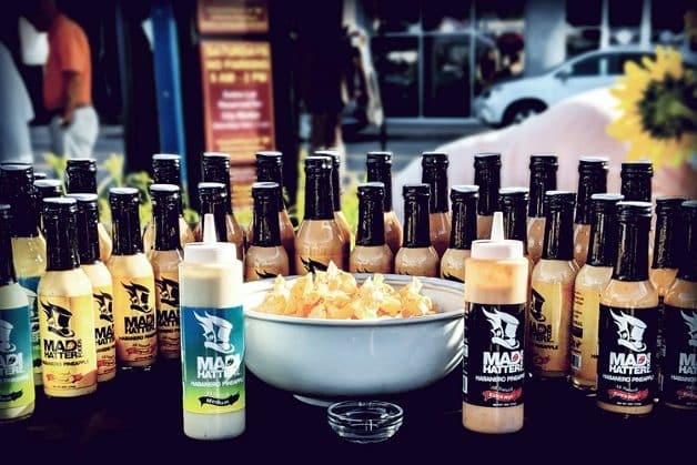 A group of bottles of Mad Hatter Hot Sauce surrounding a bowl of chips. These are made by Mad Hatter Foods and feature the hot, sweet and spicy flavors of habanero peppers and pineapple for a deliciously deviant way to celebrate Mad Hatter Day. 