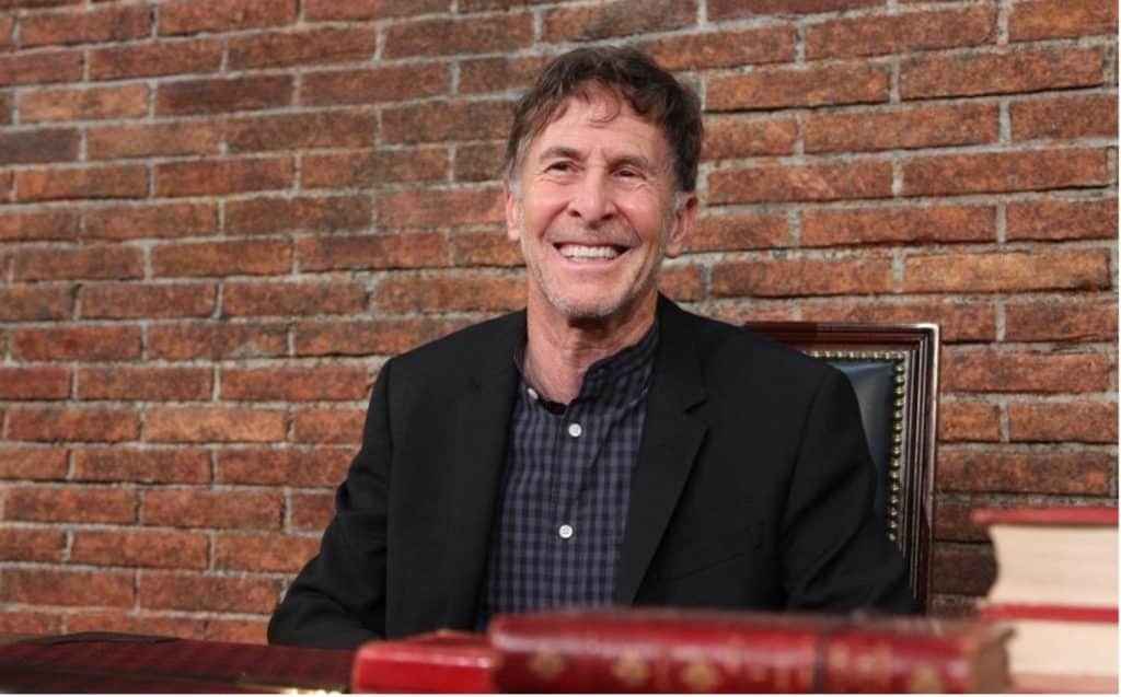 Image of Hollywood Director, Tom Schulamn, known for blockbuster films like Dead Poets Society. Here he is sitting in from of a brick wall, at a desk with some old books on it, while smiling for the camera, about to be interviewed by Author, Frank Beddor.  