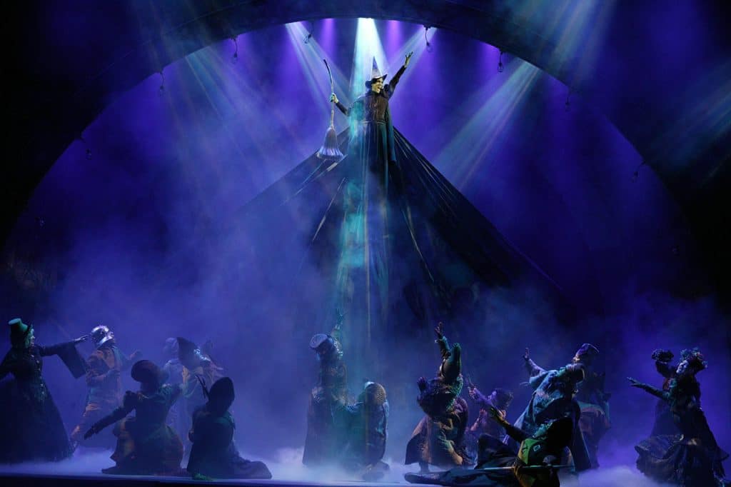 A dramatic photo from the stage production of Wicked -- Idina Menzel as the Wicked Witch Elphaba lifts into the air with dramatic blue stage lighting and the cast kneeling below in reverence