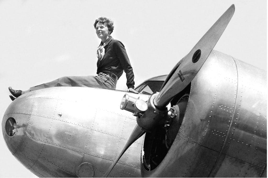 A black and white historical photograph of Amelia Earhart as she sits on the nose of her plane with the propeller in the foreground