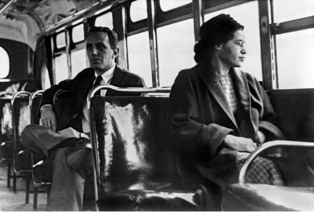 A black and white historical photograph of Rosa Parks as she sits on a bus looking out the window, a white gentleman sits in the seat row behind her.