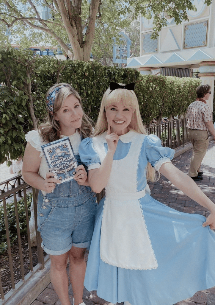 Author Sara Ella at Disneyland holding up a copy of her book, "The Wonderland Trials" next to an actress portraying Alice from Disney's 1951 film "Alice in Wonderland". 