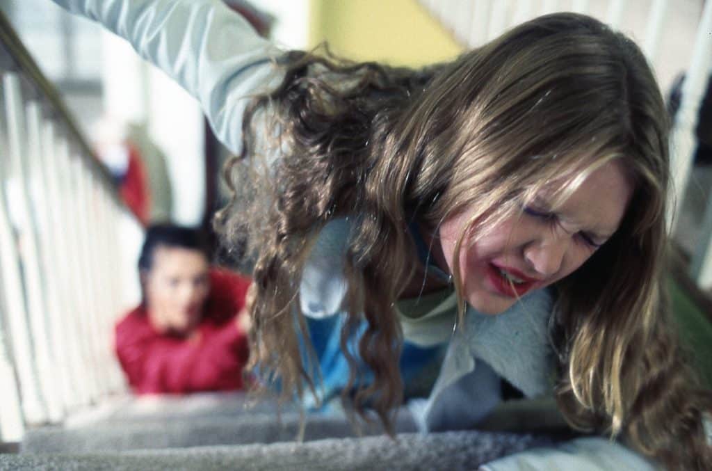 Still image from the thriller movie "Wicked" featuring Julia Stiles struggling on the stairs with Chelsea Field in the background. 
