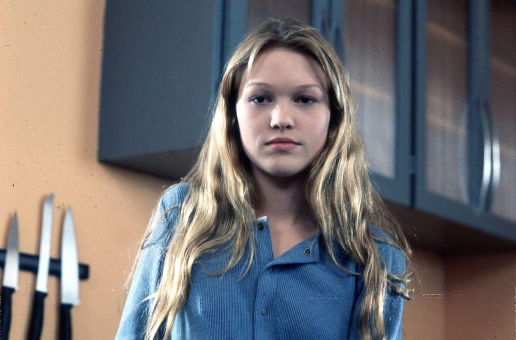 Still image from the thriller movie "Wicked" featuring Julia Stiles in a blue button-down shirt with kitchen knives hanging next to her. 