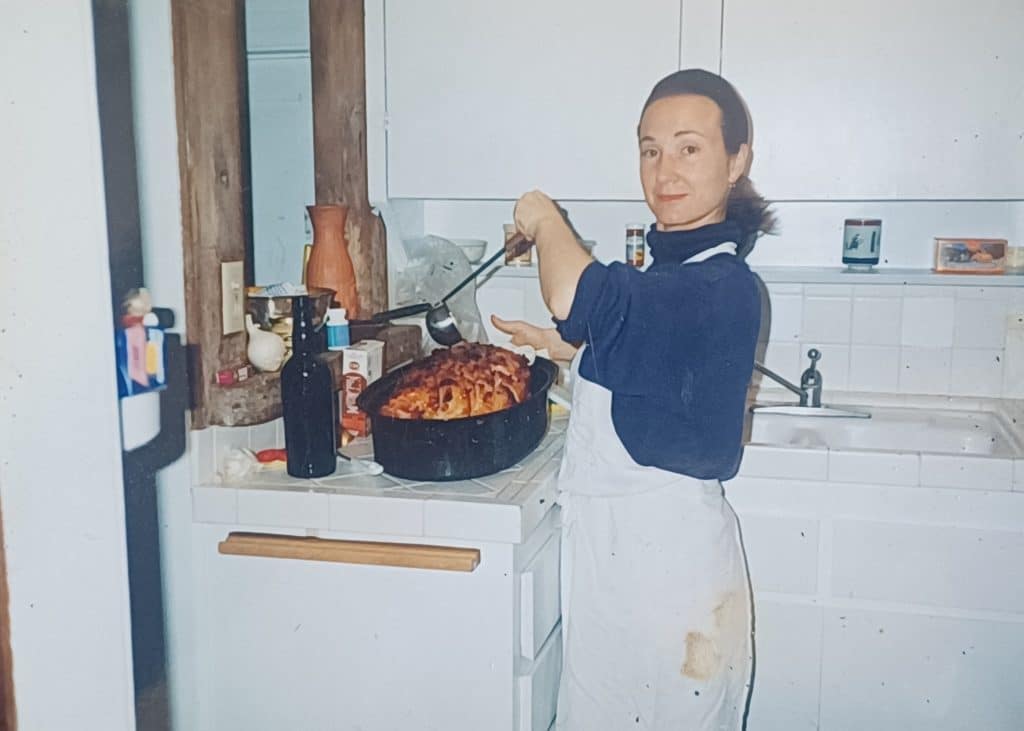 Author Liz Cavalier cooking wearing a white apron and blue turtleneck. 