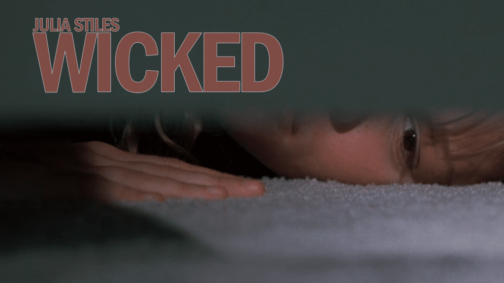 Still image from the thriller movie "Wicked" featuring Julia Stiles lying on the floor and looking underneath a door with an overlaid graphic reading "Julia Stiles" and "Wicked". 