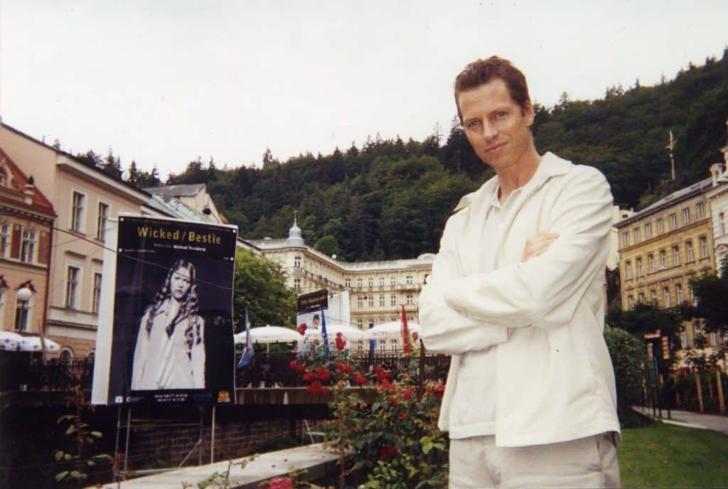 Producer Frank Beddor, dressed in white pants and a white jacket, standing next to a poster for the thriller "Wicked" at the Karlovy Vary International Film Festival in the Czech Republic. 