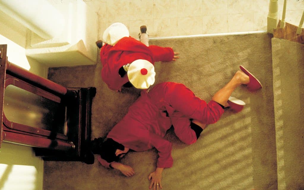 Still image from the thriller movie "Wicked" featuring Chelsea Field lying on the floor with Vanessa Zima kneeling over her, both dressed in red. 