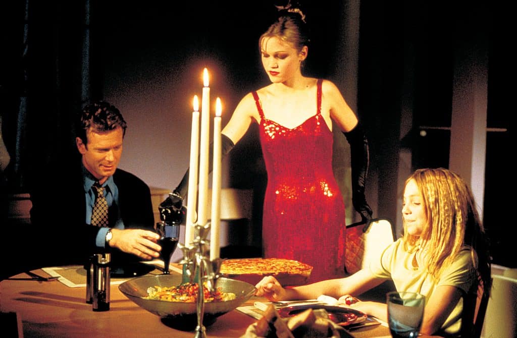 Still image from the thriller movie "Wicked" featuring Julia Stiles in a red dress pouring wine while William R. Moses and Vanessa Zima. 