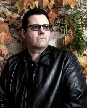 Photograph of the Edgar Award-winning Northern Irish author Adrian McKinty wearing a black leather jacket and dark tinted glasses.