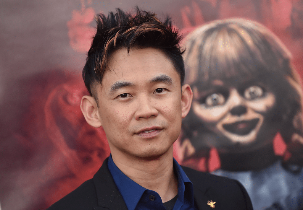 Image of "Saw" and "Furious 7" director James Wan, wearing a blue shirt and black jacket, standing in front of a promotional marquee for the 2014 supernatural horror film "Annabelle".