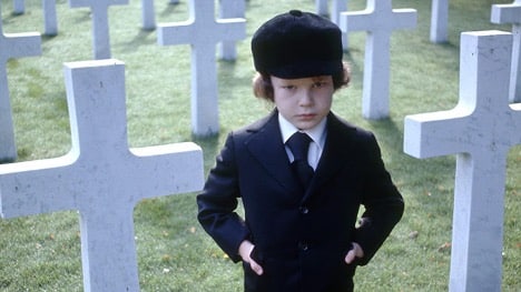 Still image from Richard Donner's 1976 supernatural horror film "The Omen," featuring Harvey Spencer Stevens as Damien Thorn in a suit and cap.