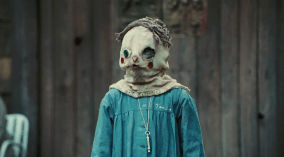 Still image of a child in a blue smock wearing a painted hood/mask from J.A. Bayona's 2007 gothic supernatural horror film "The Orphanage". 