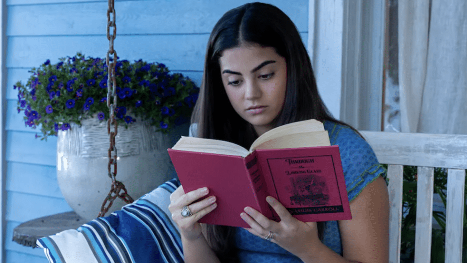 A still image from the Hallmark Channel original series "The Way Home" featuring Sadie Laflamme-Snow as Alice reading a copy of the novel "Through the Looking-Glass" by Lewis Carroll. 