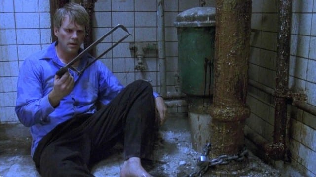Still image of Cary Elwes as Dr. Lawrence Gordon, sitting in a grungy basement staring at a saw, in the 2004 horror film "Saw".